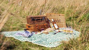 En picknickuppställning med korgar av rotting, en filt, mat och flaskor på en gräsig äng.