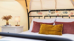 Een bed met hoofdbord versierd met kleurrijke hangende decoraties, een nachtkastje met een lamp en een vaas met gedroogde bloemen.