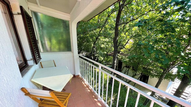 A balcony with a table and chairs, a window with blinds, and a view of trees. - Studio apartment Šarić 3 (Kraljevica)