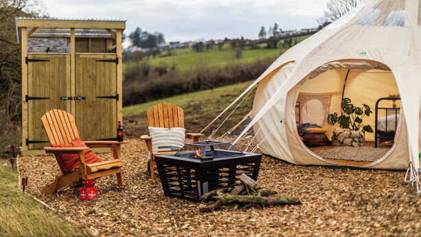 Een tent met een slaapgedeelte, een klein houten bouwwerk en twee stoelen, met een vuurkuil.