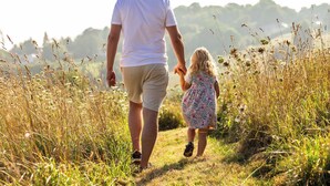 Een man en een kind wandelen hand in hand door een grasveld met hoge grassen en een heuvelachtige achtergrond.