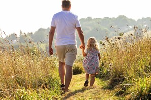 A man and a child walking hand in hand through a grassy field with tall grasses and a hilly background.