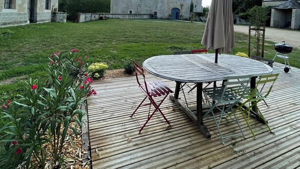 A wooden deck with a round table and chairs, a barbecue grill, and a large building in the background.