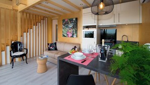 Une cuisine moderne avec des poutres en bois, un coin repas doté d’une table en verre et un espace de vie comprenant un canapé et un fauteuil.