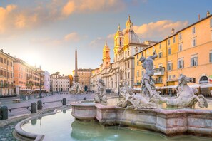 A historic fountain with classical sculptures, surrounded by classical architecture and a clear sky.