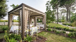 A covered outdoor seating area with tables and chairs, surrounded by a garden with various plants and trees.