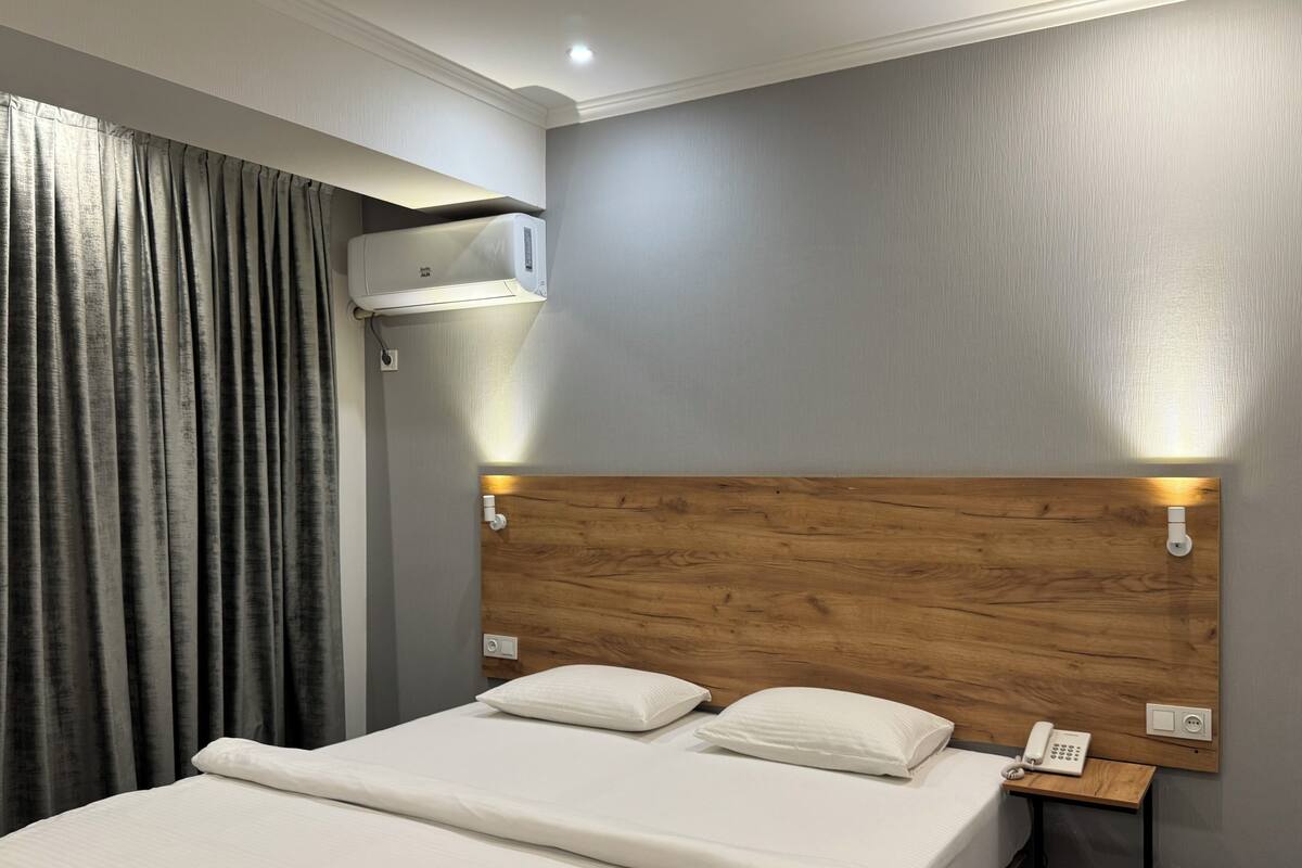 Habitación de hotel con cabecera de madera, cama con ropa de cama blanca, mesita de noche con teléfono y un equipo de aire acondicionado montado en la pared.