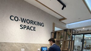 A modern co-working space with a large sign, a wooden desk, two grey chairs, and a person working on a laptop. - Afsona hotel (Qarshi)