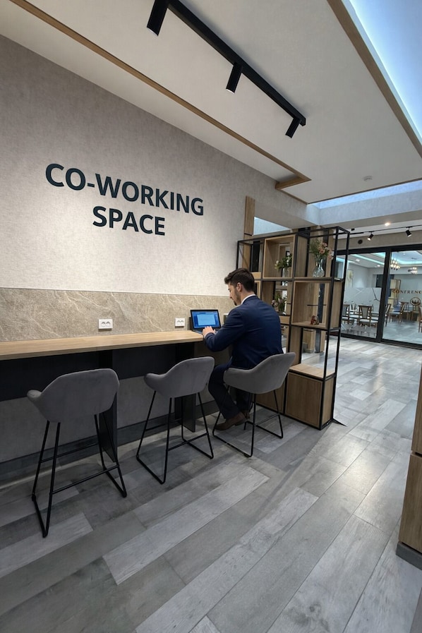 A modern co-working space with a large sign, a wooden desk, two grey chairs, and a person working on a laptop. - Afsona hotel (Qarshi)