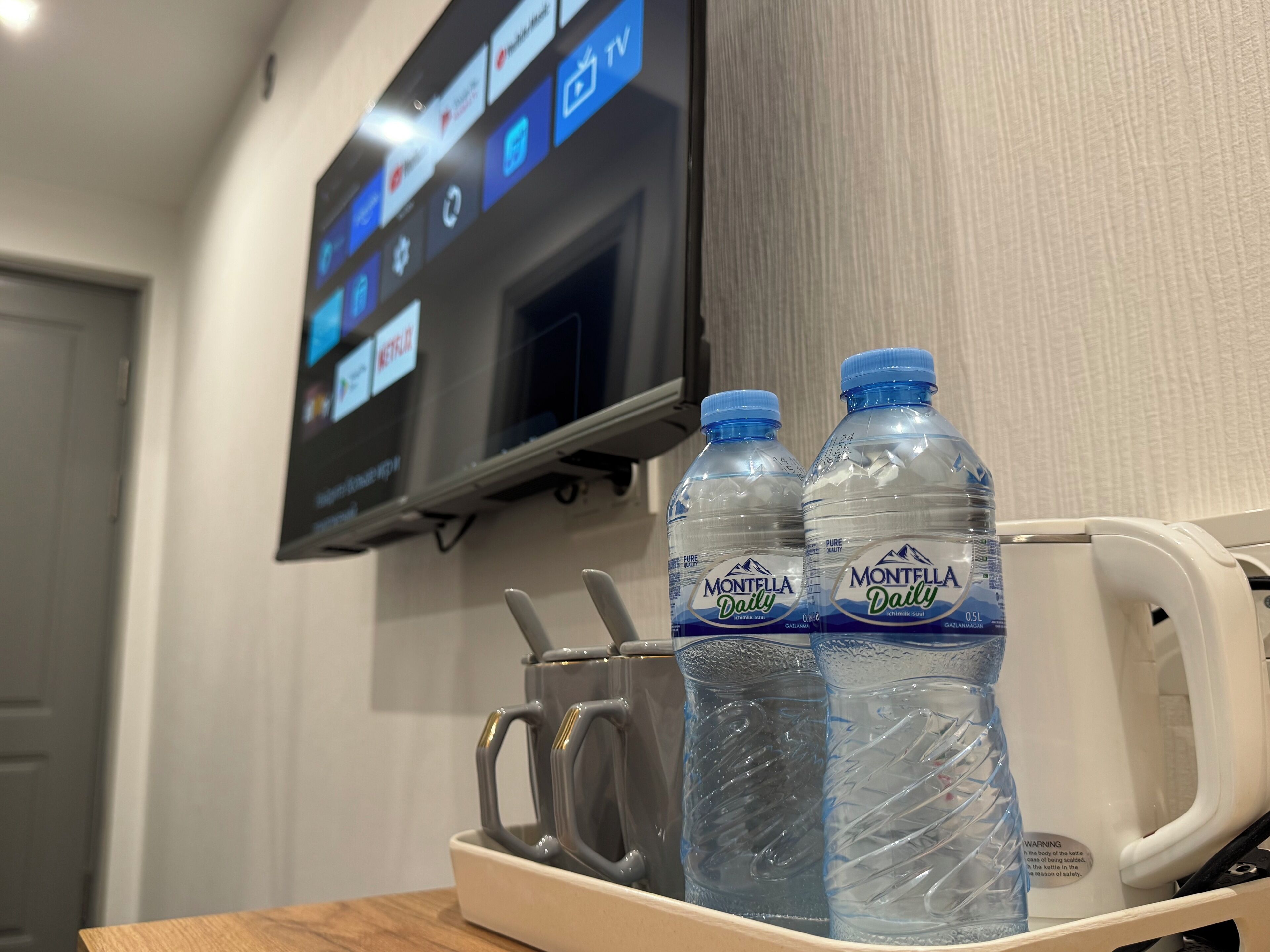 A hotel room with a TV displaying various channels, two water bottles, and a mug on a tray.