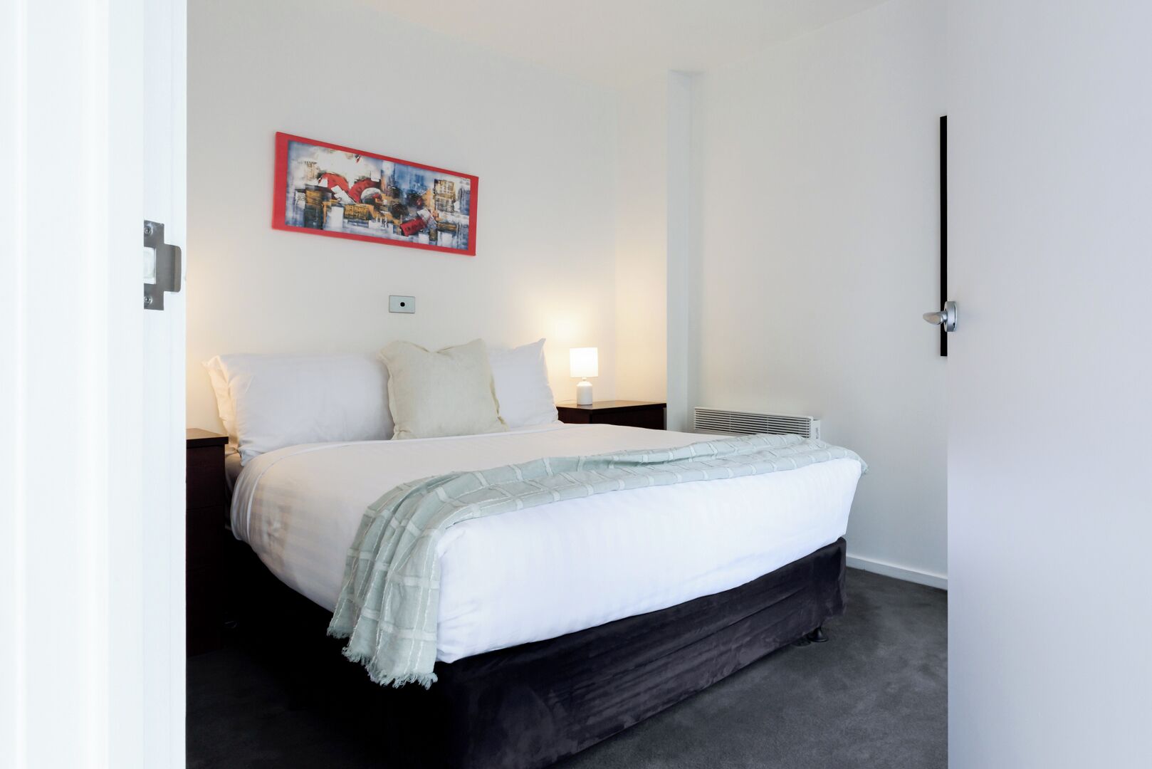 Quarto com cama, mesa de cabeceira, luminária, radiador e um quadro na parede.