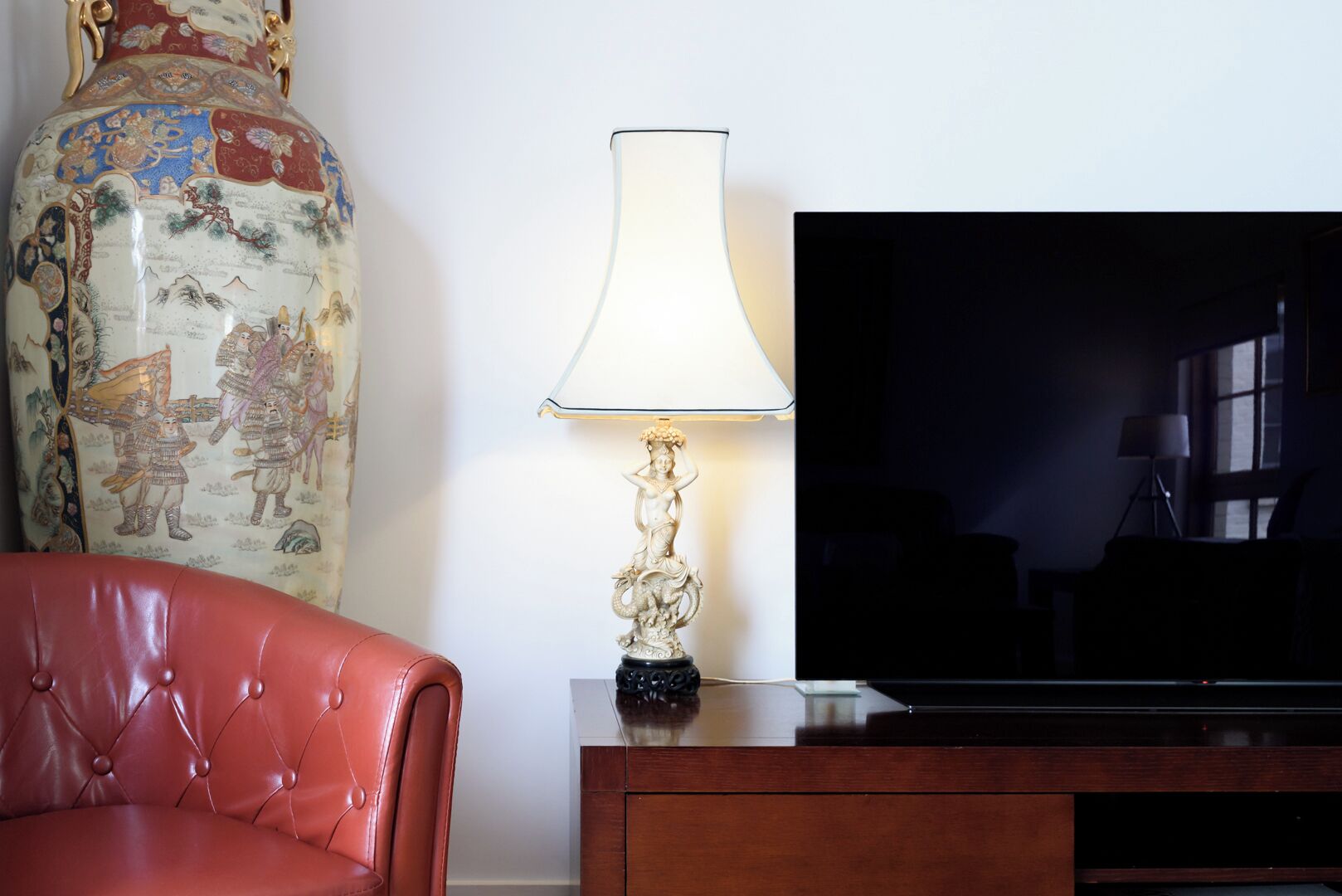 Una sala de estar con un sofá de cuero, un televisor grande sobre un soporte de madera, una lámpara decorativa y un jarrón tradicional con obras de arte.