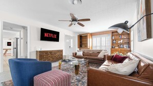 A living room with a brown leather sofa, a blue armchair, a patterned ottoman, a coffee table, and a bookshelf. A ceiling fan and a TV mounted on the wall are also visible. - Serene Pool retreat w/ Games Near Fort Moore (Columbus)