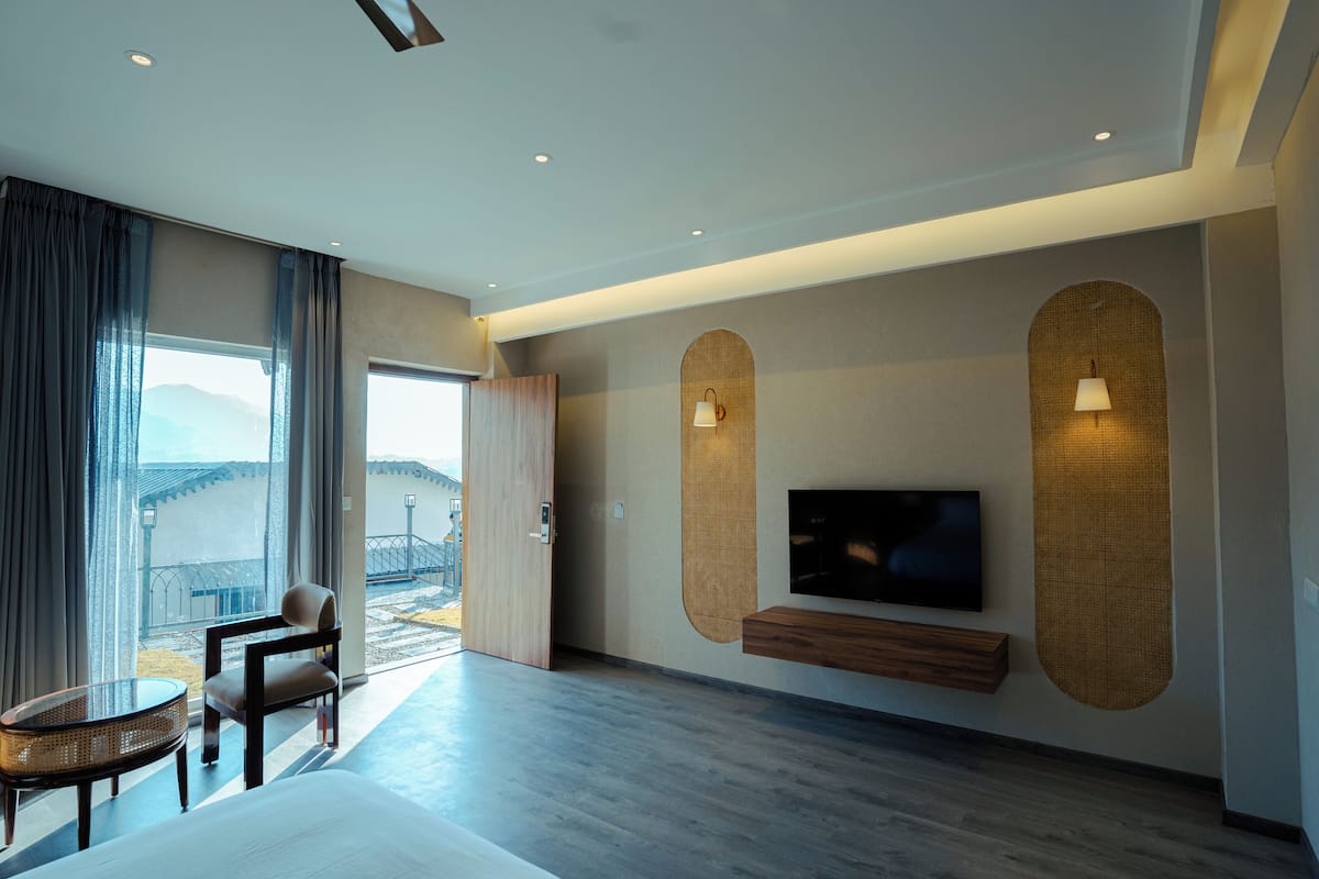 A modern hotel room with a large bed, a TV mounted on a wall, a seating area with a chair and a small table, and a view of the outdoors through a large window.