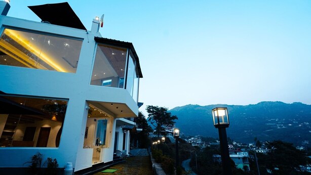 A modern two-story house with large windows and a balcony, set against a mountainous backdrop. - The Barns Resorts (Bhimtal)