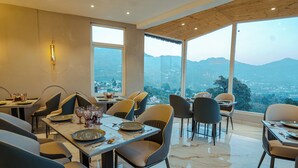 A dining area with multiple tables and chairs, large windows offering a view of the mountains. - The Barns Resorts (Bhimtal)