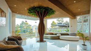 A spacious living area with a large tree planter, modern seating, and large windows offering views of the outdoors. - The Barns Resorts (Bhimtal)