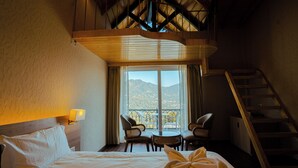 A hotel room with a bed, a small table with chairs, a balcony with a view of mountains, and a staircase. - The Barns Resorts (Bhimtal)