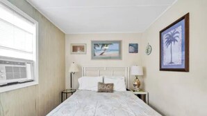 Un dormitorio con cama, una mesita de noche, una lámpara, una ventana con persianas y adornos en la pared, incluyendo cuadros enmarcados.