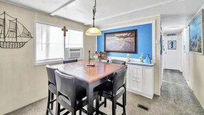 Un área de comedor con una mesa y sillas de madera, una lámpara colgante, un cuadro enmarcado en la pared y un armario blanco con un detalle en pared azul.