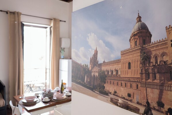Uma área de refeições com uma mesa posta para dois, uma janela com cortinas e uma grande moldura com a imagem de um edifício histórico.