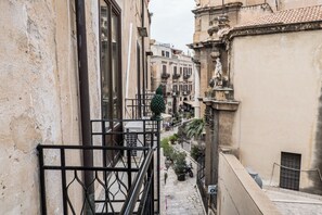 Uma rua estreita com edifícios em ambos os lados, uma pequena varanda com grades e uma estátua à direita.