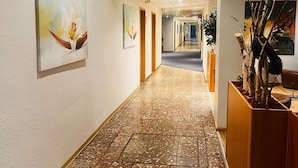 A hallway with a terrazzo floor, framed artwork on the walls, and a wooden reception desk with a plant. - Werrapark Hotel Heubacher Höhe (Masserberg)