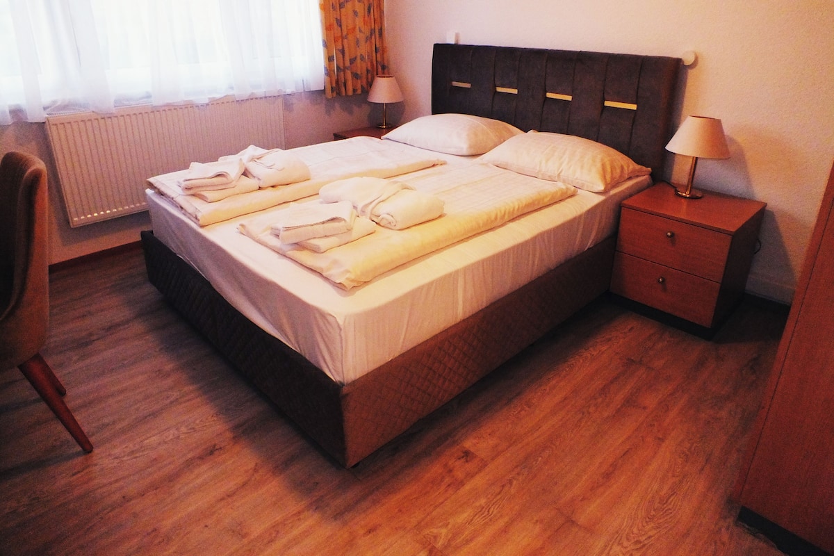 Een opgemaakt bed met handdoeken, een nachtkastje met een lamp en een stoel in een hotelkamer.