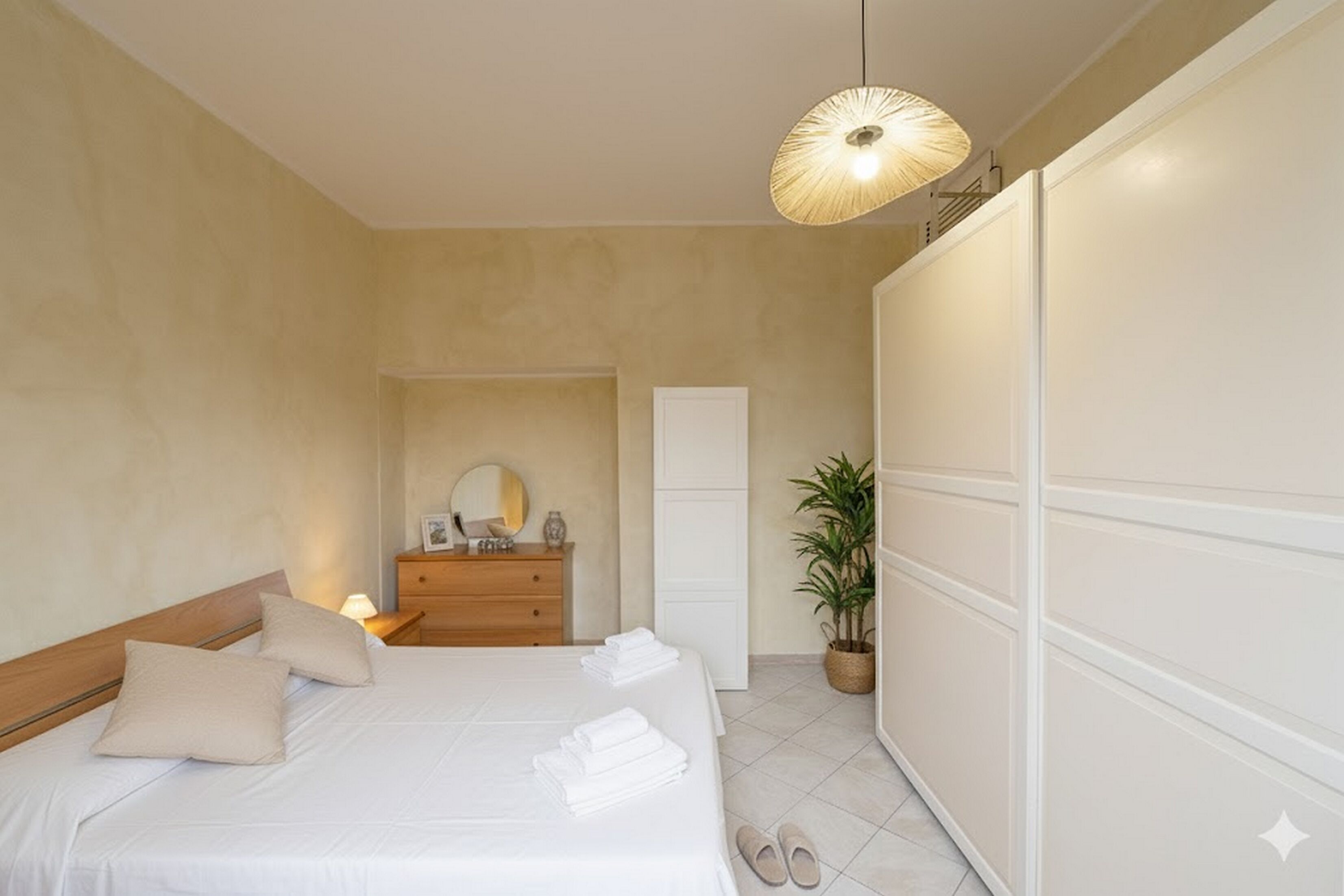Un dormitorio con cama, mesita de noche, lámpara, cómoda, espejo, planta en maceta y armario.