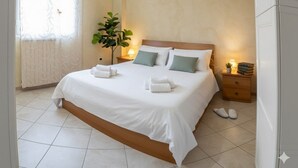 A hotel room with a large bed, two bedside tables, a potted plant, and a window with curtains. - Littardi Smart Suite (Imperia)