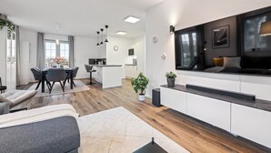A modern living room with a sofa, a dining area with a table and chairs, a kitchen area, and a large TV on the wall.