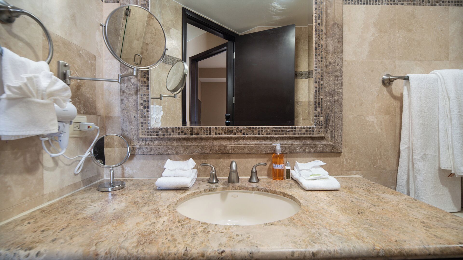 A bathroom with a granite countertop, a sink, three faucets, two mirrors, and a bottle of soap.