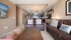 Ein geräumiges Wohnzimmer mit einem braunen Ledersofa, einem Holztisch und bunten, gewebten Poufs. Im Hintergrund ist eine Küche mit modernen Geräten zu sehen.