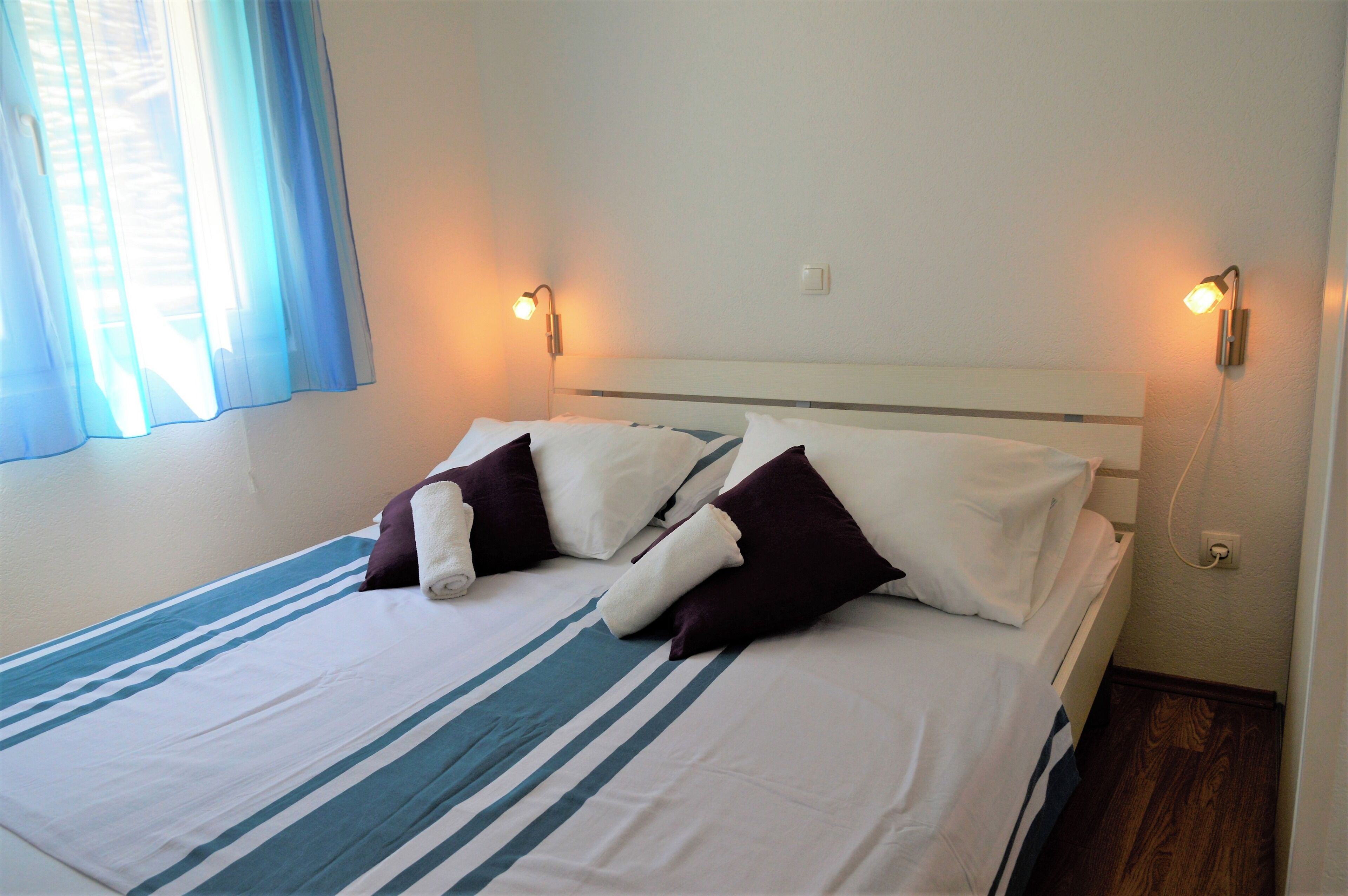 Una cama bien tendida con ropa de cama a rayas y dos almohadas, dos luces nocturnas fijadas en la pared y una ventana con cortinas azules.