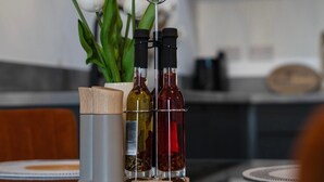 Eine Küchenarbeitsplatte mit einer Olivenölflasche, einer roten Weinflasche und einer weißen Weinflasche. Im Hintergrund steht eine Pflanze mit weißen Blüten.