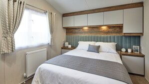 A neatly made bed with a grey blanket, wooden headboard, and bedside tables with clocks and picture frames.