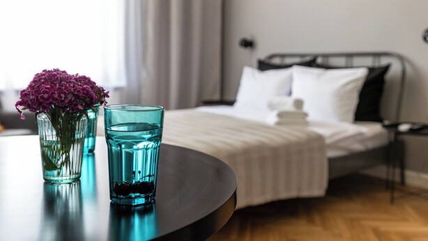 Une chambre d’hôtel avec un lit, une table de chevet ornée d’un vase de fleurs et d’un verre d’eau, et une vue sur l’extérieur.