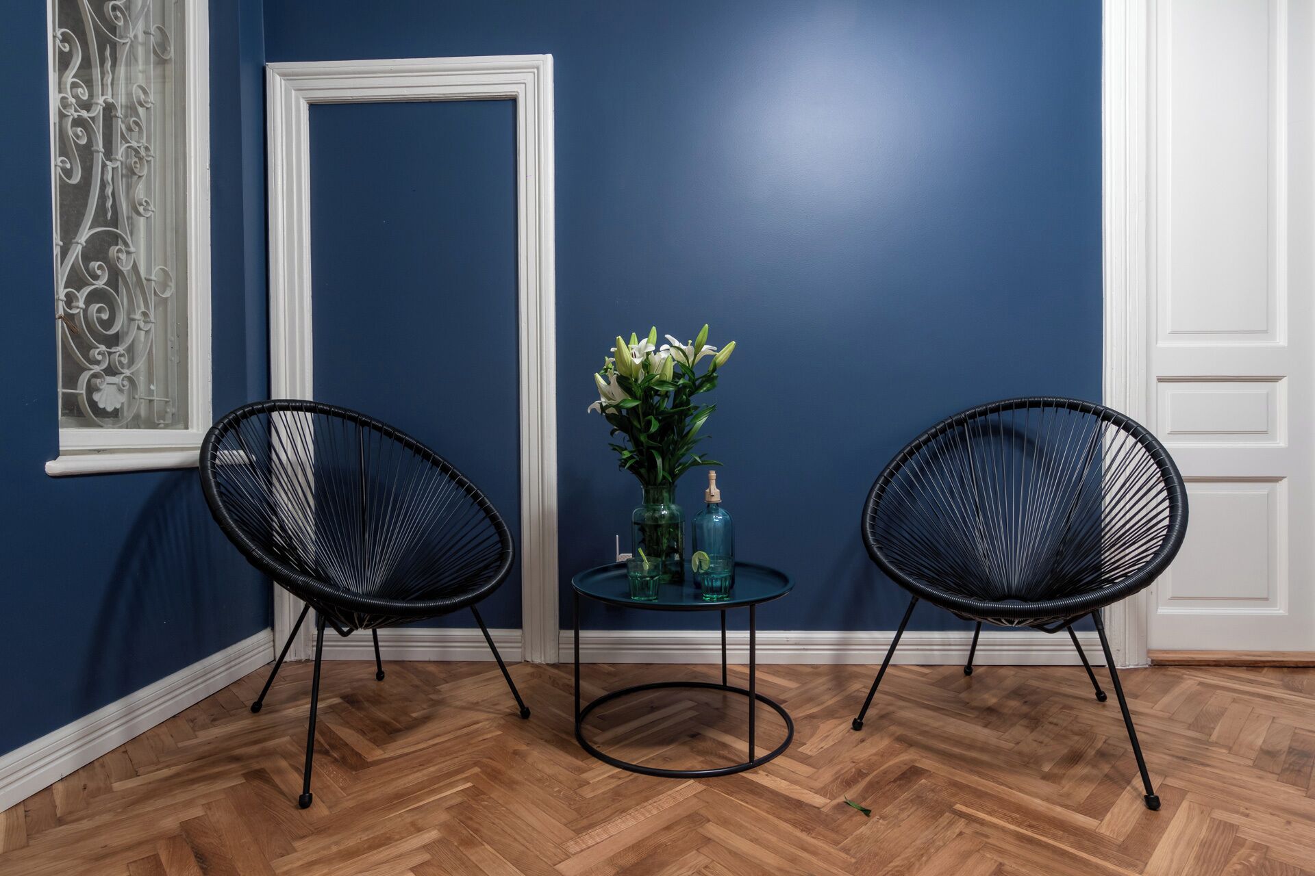 A room with two black wireframe chairs, a small round table with a vase of flowers, and a large mirror on the wall.