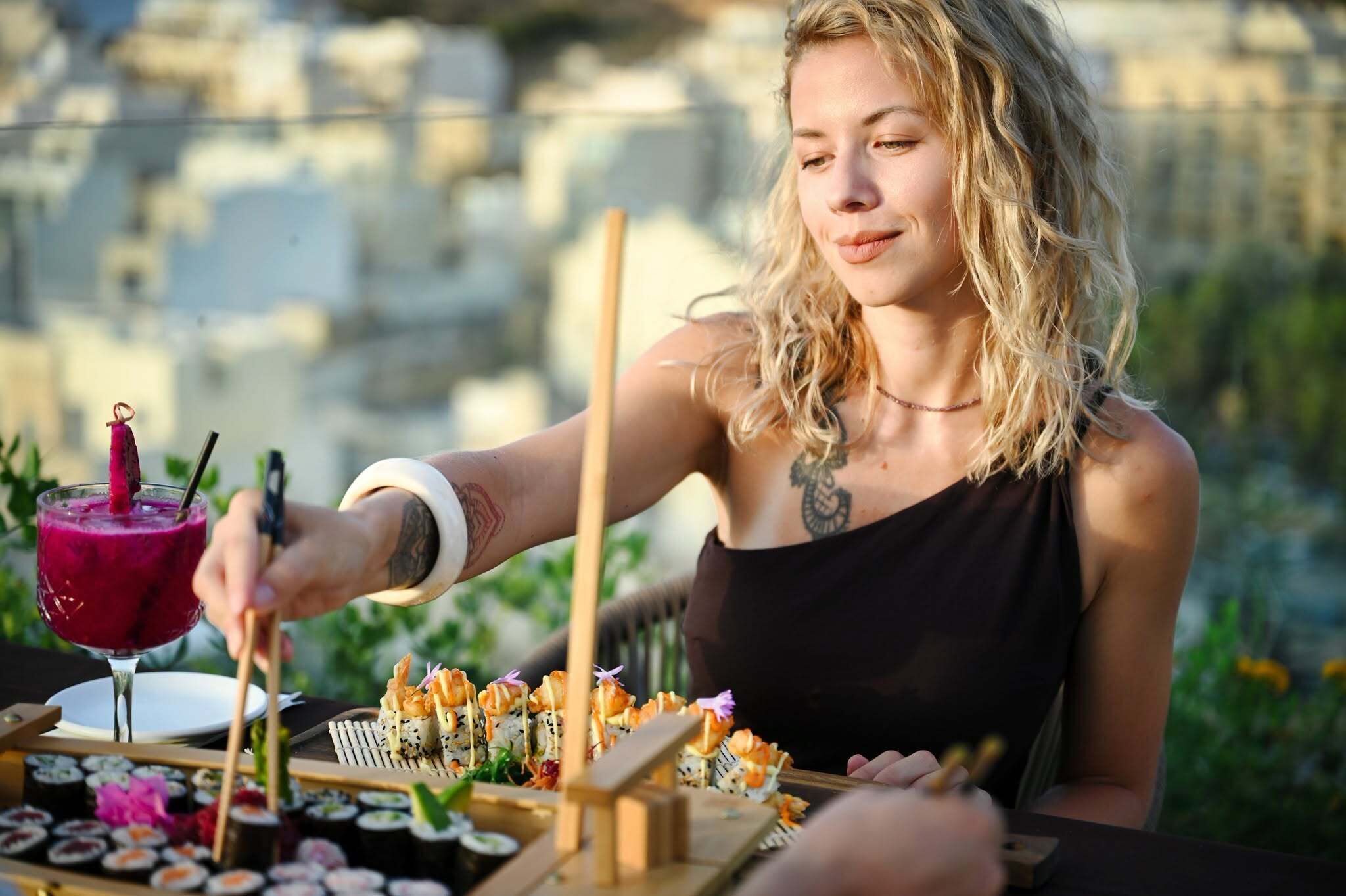 A person serving sushi at an outdoor dining area with a cityscape in the background.