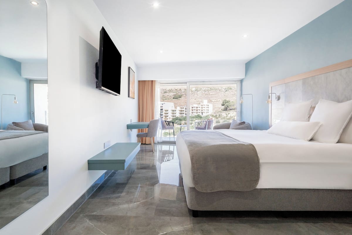 Quarto de hotel moderno com uma cama grande, uma TV fixada na parede, uma mesa de trabalho e vista para prédios pela janela.