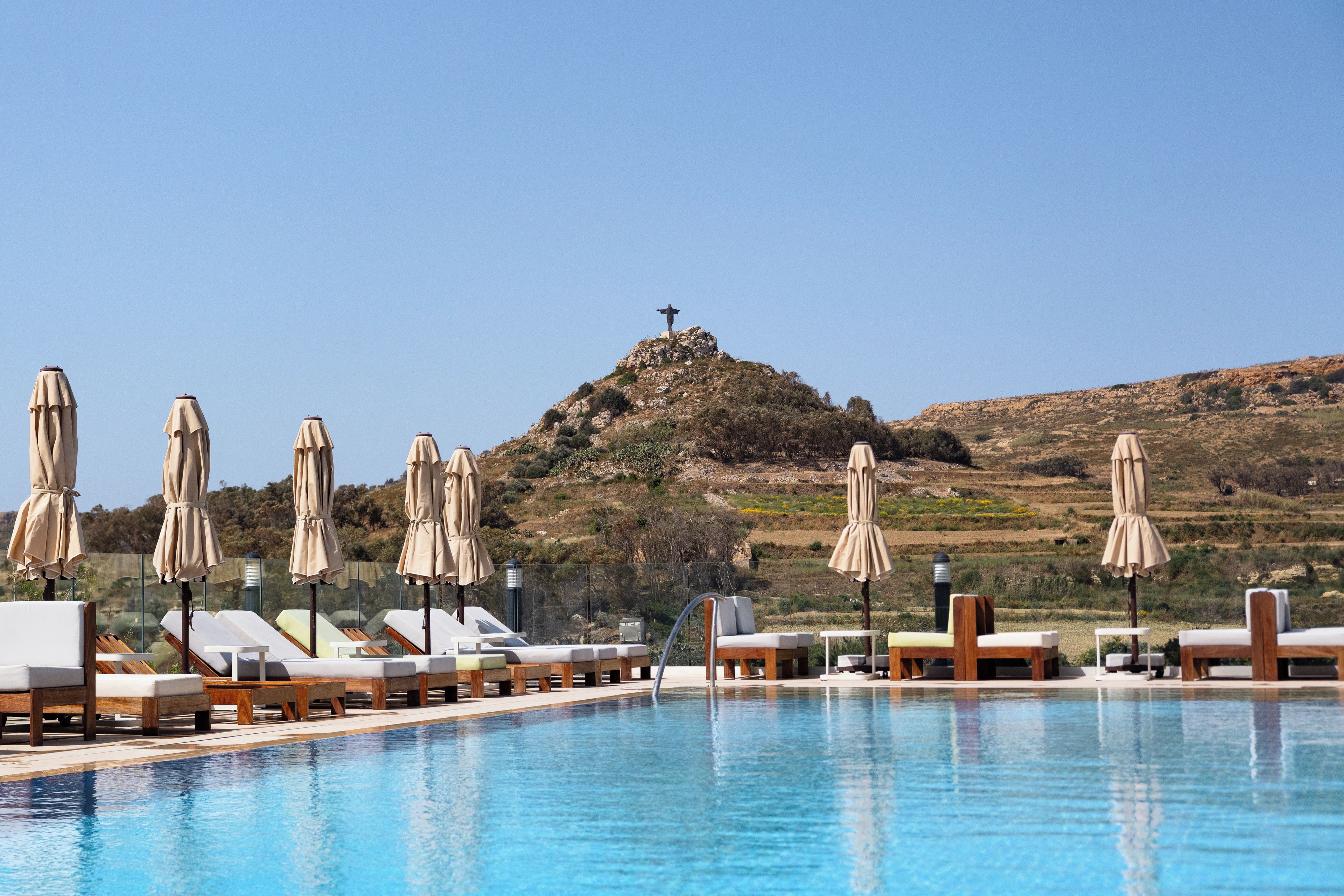 Ein Poolbereich mit Liegestühlen und Sonnenschirmen, eine Statue auf einem Hügel und ein klarer Himmel.