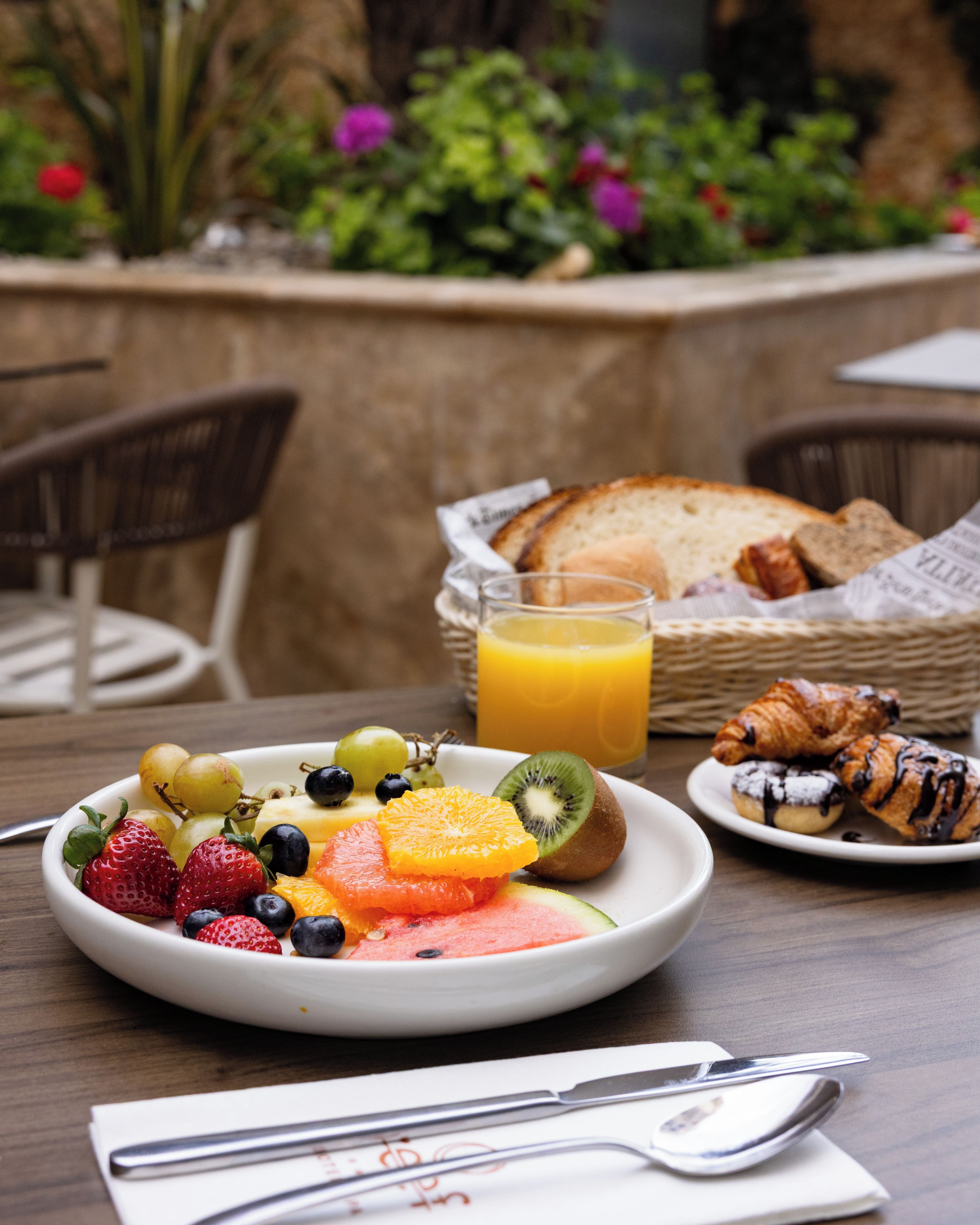 Un desayuno con una fuente de fruta, un vaso de zumo de naranja y cruasanes sobre una mesa.