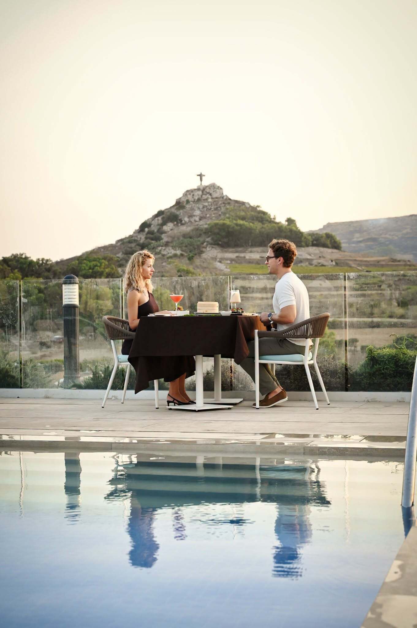 A dining table with chairs, a pool, and a view of a hill with a cross.