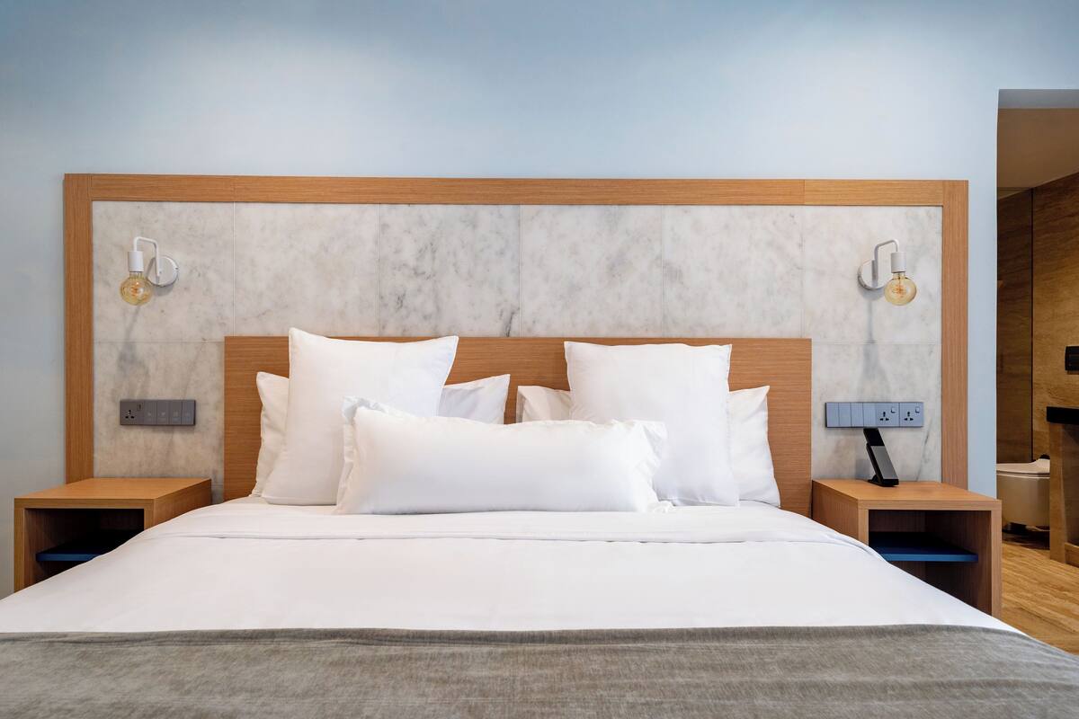 Cama bem arrumada com lençóis e almofadas brancos, cabeceira de madeira, mesas de cabeceira com abajures e uma tomada na parede.