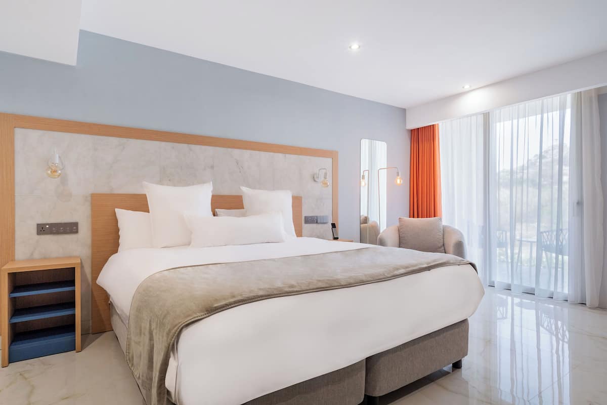 Quarto de hotel moderno com uma cama grande, um pequeno espaço de estar e uma janela ampla com cortinas.