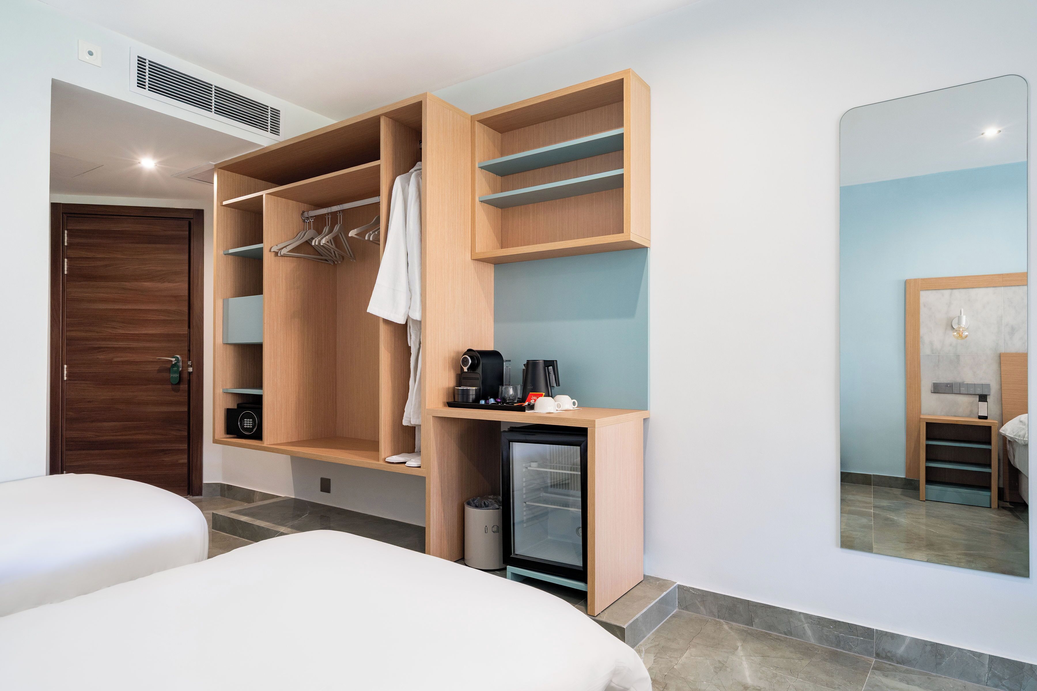 A hotel room with a wooden wardrobe, a small refrigerator, a mirror, and two beds.