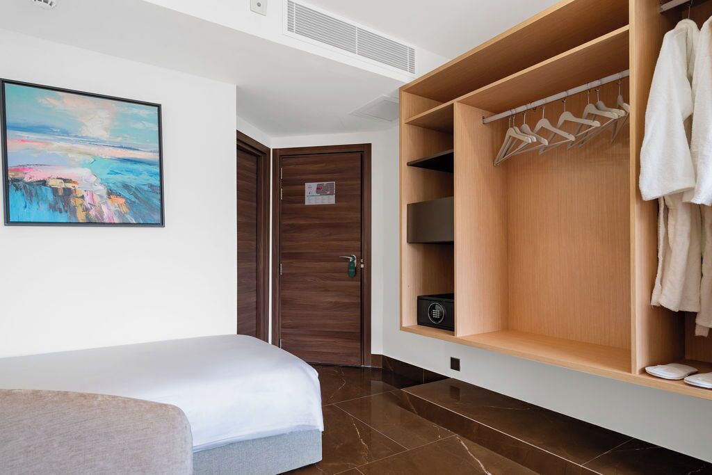 A hotel room with a bed, a wooden wardrobe with hangers, a painting on the wall, and a door.