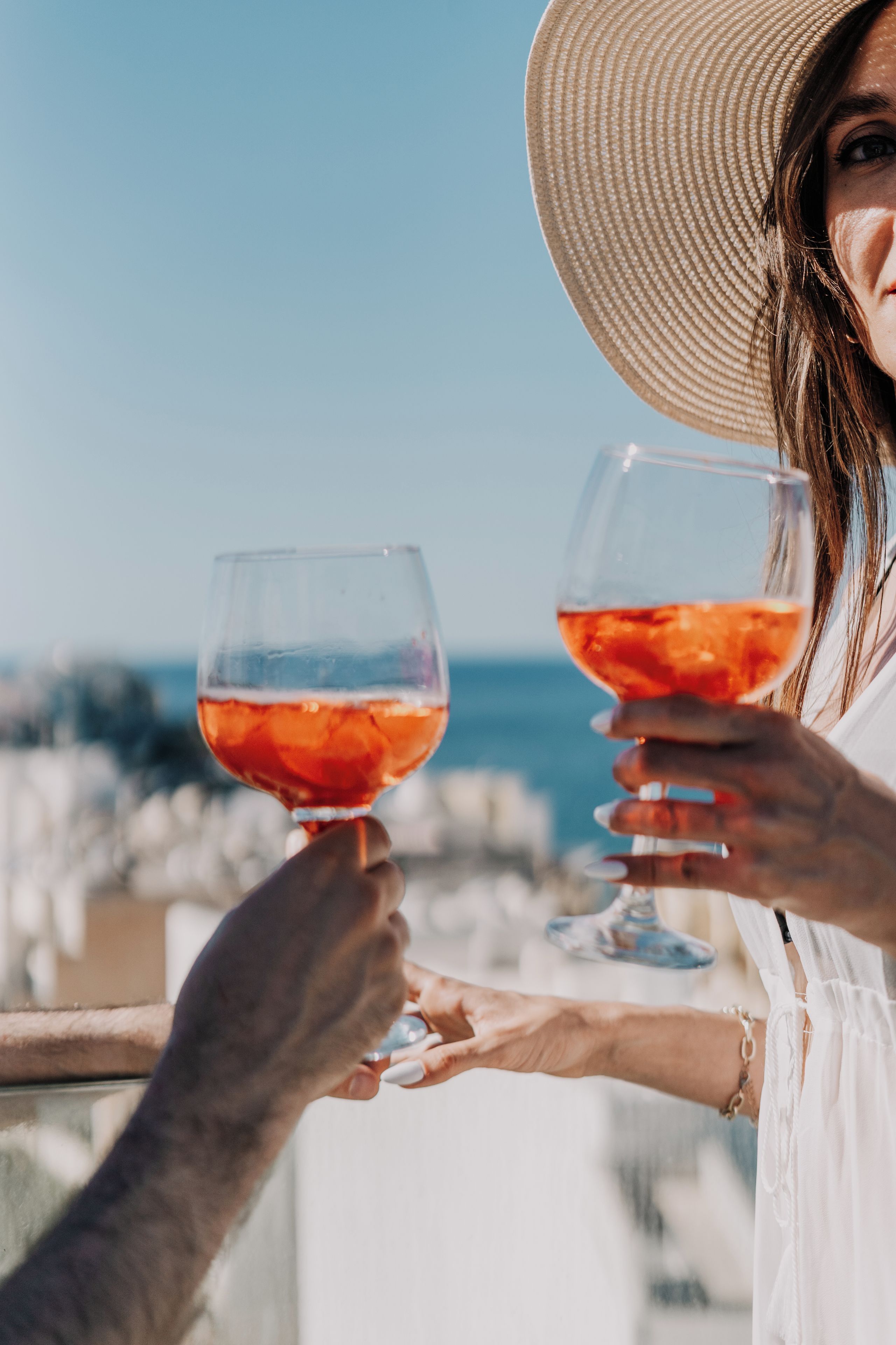 Two people holding wine glasses with a clear liquid, possibly a cocktail, against a backdrop of a clear sky and a body of water.