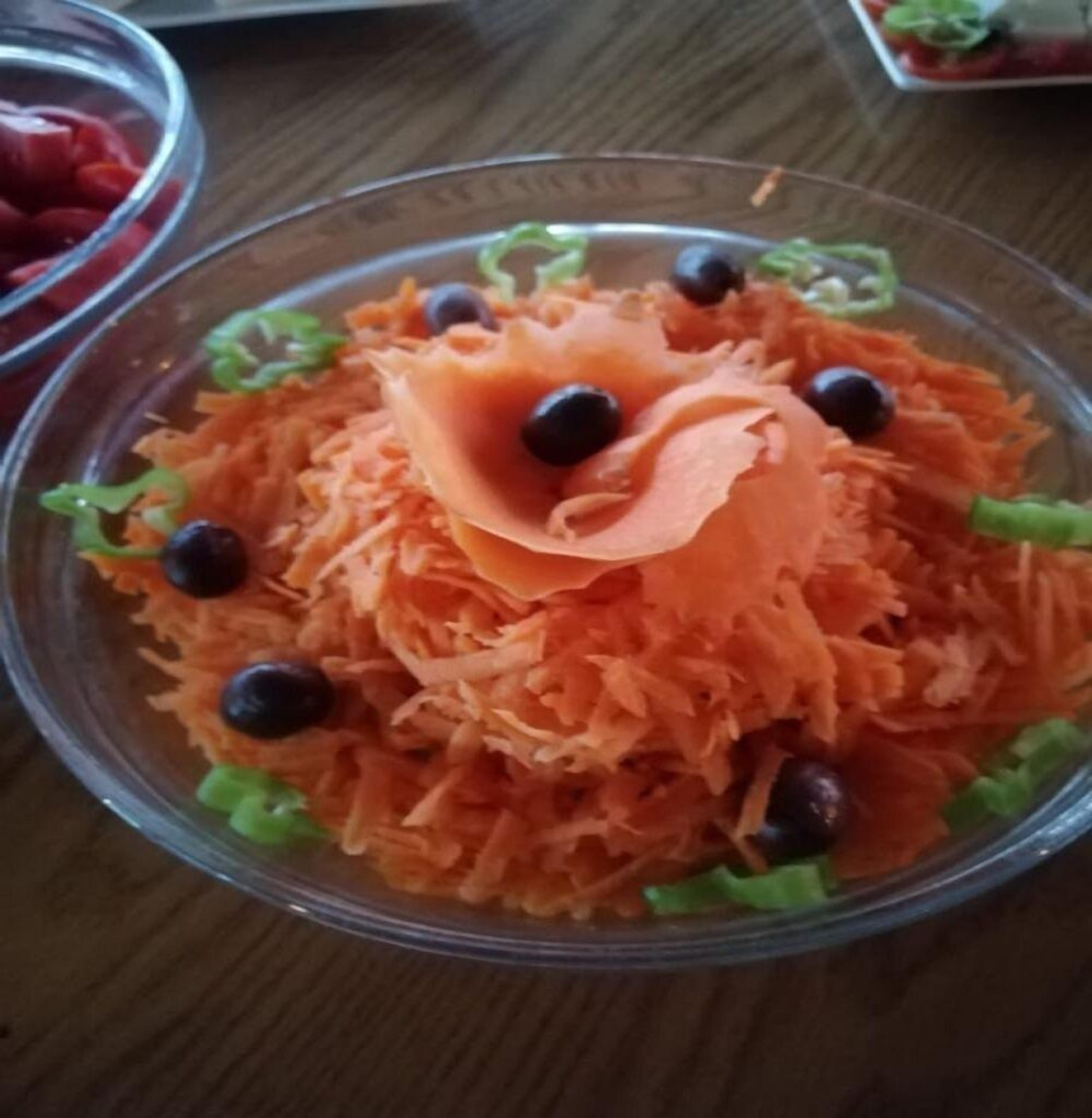 A clear glass bowl containing shredded carrots, green lettuce, and black olives.