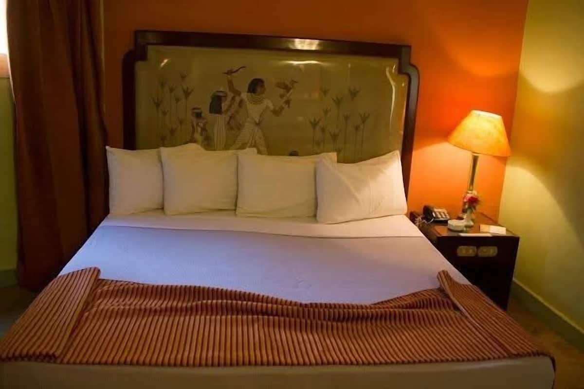 A hotel room with a bed, pillows, a striped blanket, a nightstand with a lamp, and a wall with a decorative panel.